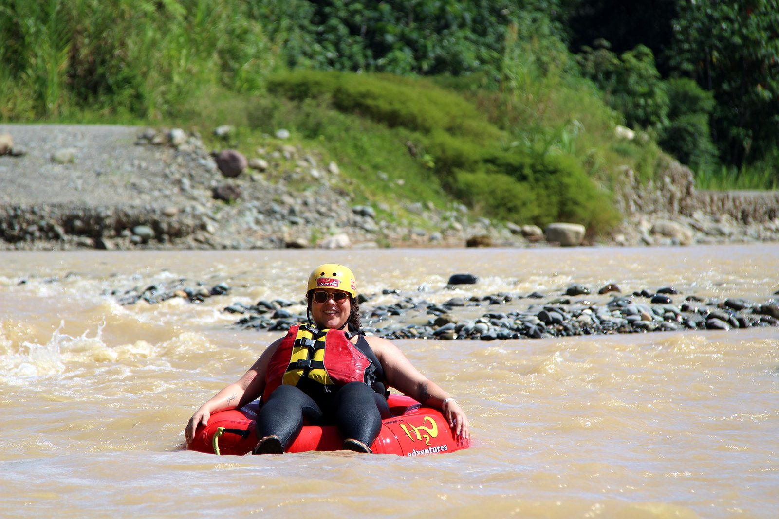 A smiling guest sits in her red tube in front of a gravel bar